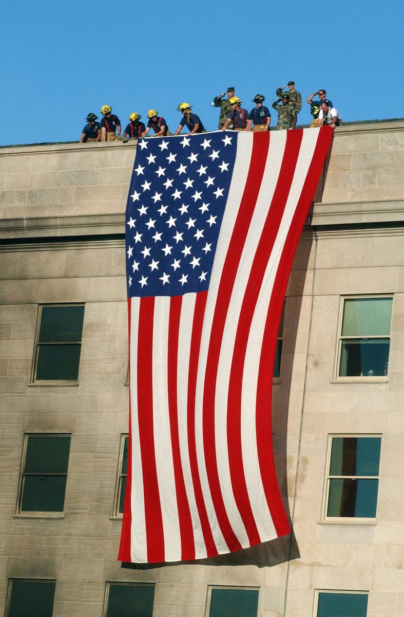 The original finding aid described this photograph as: [Complete] Scene Caption: Soldiers from the 3rd Infantry render honors as fire fighters and rescue workers unfurl a huge American flag over the side of the Pentagon as rescue and recovery efforts continued following the September 11, 2001 terrorist attack. The flag, a garrison flag, sent from the US Army Band at nearby Fort Myer, Virginia, is the largest authorized (20 x 38) flag for the military. Shortly after 8 AM on September 11, 2001 in an attempt to frighten the American people, five members of Al-Qaida, a group of fundamentalist Islamic Muslims, hijacked American Airlines Flight 77, a Boeing 757-200, from Dulles International Airport just outside Washington DC. About 9:30 AM they flew the aircraft and 64 passengers into the side of the Pentagon. The impact destroyed or damaged four of the five 
