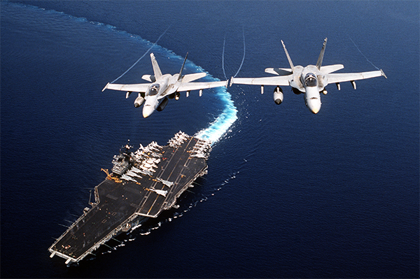 Aerial view two F/A-18C Hornet aircraft of Strike Fighter Squadron Seventy-Four (VFA-74) in flight above the Forrestal Class, Aircraft Carrier USS SARATOGA (CV 60) during Operation Desert Shield. The SARATOGA is making a hard turn to starboard and is in the background. Two F/A-18C Hornet aircraft of Strike Fighter Squadron 74 fly above the Forrestal-class aircraft carrier Saratoga (CV-60) making a hard turn to starboard during Desert Shield, 4 November 1990. (U.S. Navy photo, 91-159-H)